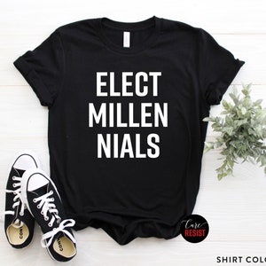 May include: Black t-shirt with white text that reads "ELECT MILLENIALS". The shirt is on a white wooden surface with a small green plant and a pair of white sneakers.