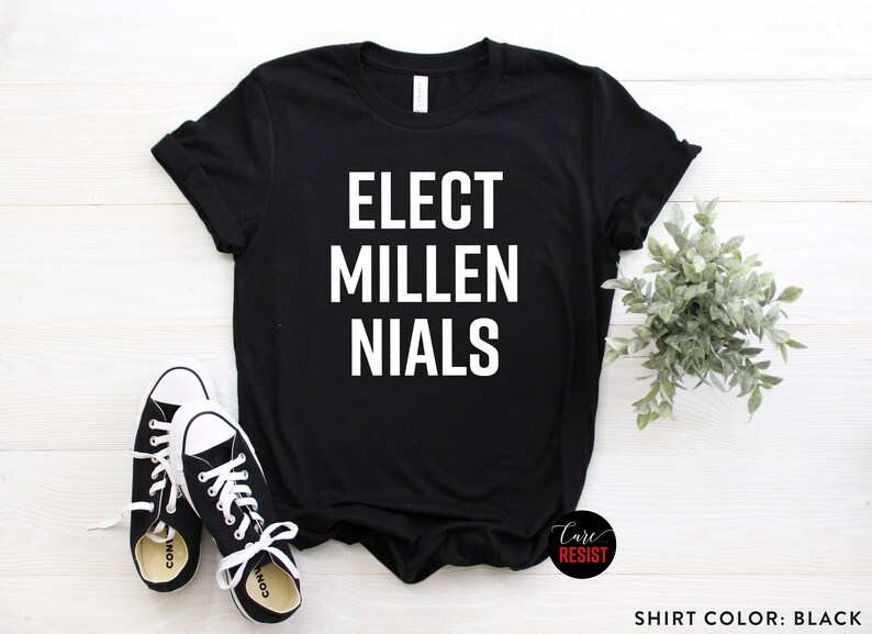 May include: Black t-shirt with white text that reads "ELECT MILLENIALS". The shirt is on a white wooden surface with a small green plant and a pair of white sneakers.