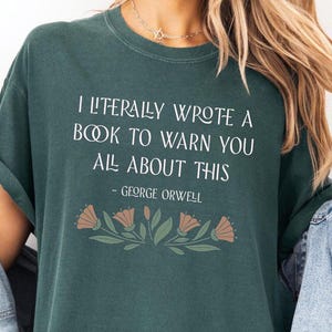 May include: A dark green t-shirt with a white graphic print that reads "I literally wrote a book to warn you all about this - George Orwell". The graphic is surrounded by a floral design in shades of green and peach.