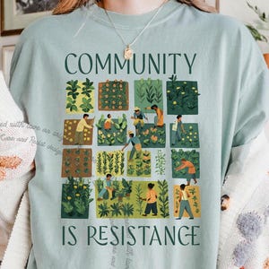 Könnte beinhalten: Ein hellblaues T-Shirt mit einem grafischen Design eines Gemeinschaftsgartens. Die Grafik zeigt Menschen mit unterschiedlichen Hautfarben, die Pflanzen in einem Raster von Quadraten pflegen. Der Text "COMMUNITY IS RESISTANCE" ist unter der Grafik gedruckt.