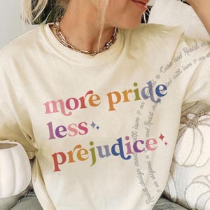 May include: Cream-coloured sweatshirt featuring the phrase "more pride less prejudice" in rainbow hues. The design includes a star and diamond. A gold chain necklace is visible at the neckline.