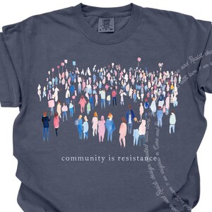 May include: A dark blue t-shirt with a watercolor illustration of a crowd of people holding signs. The text "community is resistance" is printed below the illustration.