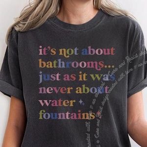 May include: A dark grey t-shirt with a colourful graphic that reads "it's not about bathrooms... just as it was never about water + fountains".