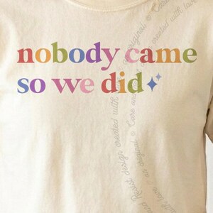 May include: A cream-colored t-shirt featuring the multicolored text "nobody came so we did" printed in a serif font across the chest. The design includes a small four-pointed star icon at the end of the text, rendered in a muted blue shade.