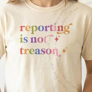 May include: A cream-coloured short-sleeve t-shirt featuring the text "reporting is not treason" printed in a multi-coloured, retro-style font. The design includes small star-shaped accents to the right of the text and a faint watermark.