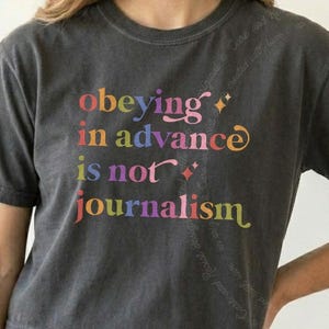 Puede incluir: Una camiseta de manga corta gris carbón con el texto multicolor "obeying in advance is not journalism" impreso en la parte delantera. Las letras tienen una fuente decorativa con pequeñas estrellas intercaladas en todo el diseño.