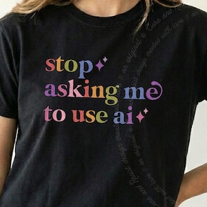 May include: A black short-sleeved t-shirt featuring the multicoloured text "stop asking me to use ai" printed on the front. The design includes small four-pointed star icons interspersed among the words, which are rendered in various vibrant hues.