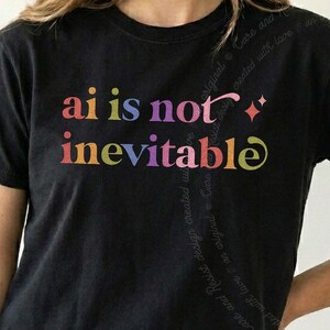 Peut inclure: Un t-shirt noir à manches courtes avec le texte multicolore "ai is not inevitable" imprimé sur la poitrine dans une police stylisée. Le motif comprend une petite icône d'étoile à quatre branches à droite du texte, sur un fond en tissu noir uni.