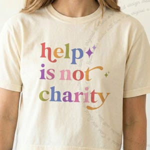 May include: A cream-colored short-sleeve t-shirt featuring the multi-colored text "help is not charity" in a retro font. The design includes small star accents and faint, cursive text flowing diagonally across the right side of the shirt.