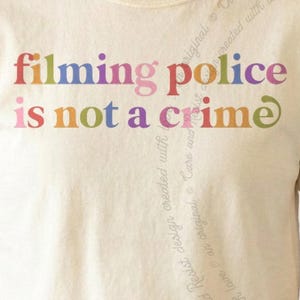 May include: A cream-coloured t-shirt featuring the text "filming police is not a crime" printed in a variety of vibrant, multi-coloured letters. The words are arranged in two lines across the chest, with each letter displaying a different hue.