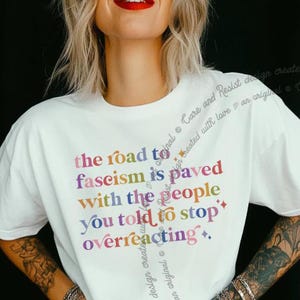Puede incluir: Camiseta blanca con un estampado gráfico colorido que dice "El camino al fascismo está pavimentado con la gente a la que le dijiste que dejara de reaccionar en exceso."