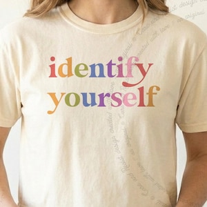 May include: A cream-colored short-sleeve t-shirt featuring the text "identify yourself" printed in a multi-colored gradient font across the chest. The letters appear in shades of red, orange, yellow, green, blue, purple, and pink.