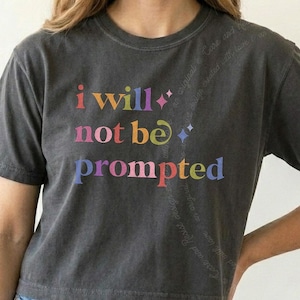Puede incluir: Una camiseta de manga corta en color gris carbón con el texto multicolor "I will not be prompted" impreso en el frente. El diseño incluye pequeños detalles en forma de estrella y una marca de agua diagonal sutil en el lado derecho.