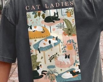 May include: A dark gray t-shirt with a colorful illustration of cats lounging in a room with plants. The text "CAT LADIES FOR KAMALA" is printed on the shirt.