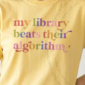 Może przedstawiać: Bladożółty t-shirt z napisem "my library beats their algorithm" wydrukowanym wielokolorową czcionką szeryfową. Litery są w odcieniach czerwieni, pomarańczu, żółci, zieleni, błękitu i fioletu, z małą ikoną czteroramiennej gwiazdki.