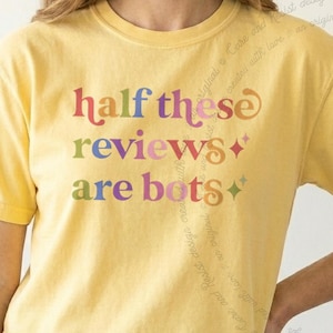 May include: A yellow short-sleeve t-shirt featuring the multi-colored text "half these reviews are bots" printed on the front. The lettering uses a stylized, rounded font in shades of red, blue, purple, orange, and green, accompanied by two small four-pointed star icons.