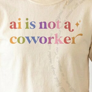 May include: A cream-colored t-shirt featuring the multicolored text "ai is not a coworker" printed across the chest in a retro-inspired font. The design includes small star accents and a faint, diagonal watermark of repeating text.