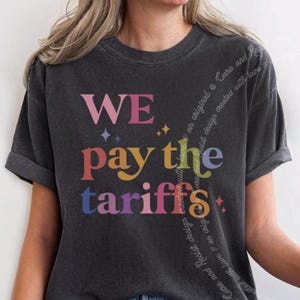 Puede incluir: Una camiseta gris oscuro con el texto "WE pay the tariffs" en colores arcoíris. El texto está estilizado con un efecto de brillo.