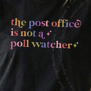 May include: A black t-shirt featuring the multi-colored text "the post office is not a poll watcher" printed in a stylized font. The design includes small four-pointed star icons interspersed among the words in shades of pink, blue, and yellow.