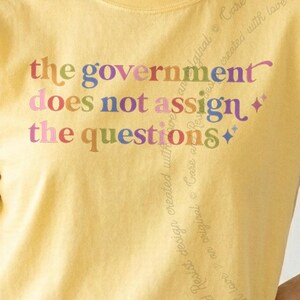 May include: A pale yellow t-shirt featuring the text "the government does not assign the questions" printed in a multi-coloured, serif font. The design includes small four-pointed star icons on the right side of the text in a soft colour palette.
