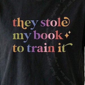 Op de afbeelding: Een zwart t-shirt met de meerkleurige tekst "they stole my book to train it" gedrukt in een schreeflettertype. De letters variëren in kleuren zoals oranje, geel, groen, blauw, paars en roze, met twee kleine vierpuntige sterren.