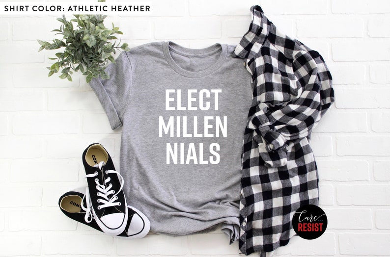 May include: A heather grey t-shirt with the text "ELECT MILLENNIALS" printed in white. The shirt is laid flat on a white brick background with a plaid shirt and black sneakers.