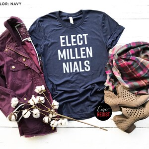 May include: A navy blue t-shirt with the text "Elect Millenials" in white lettering. The shirt is laid flat on a white wooden surface with a burgundy jacket, a plaid scarf, and two pairs of brown ankle boots.