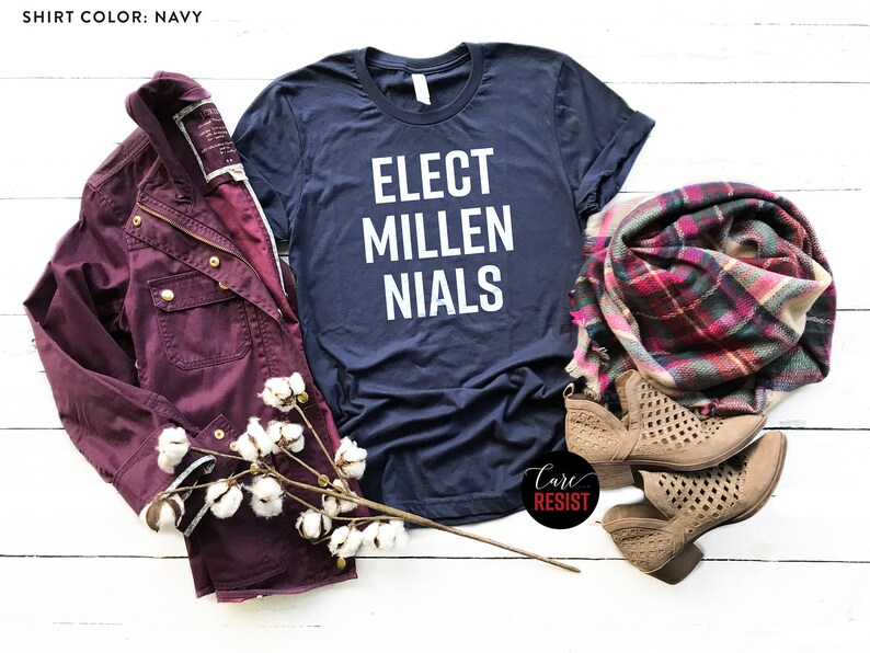 May include: A navy blue t-shirt with the text "Elect Millenials" in white lettering. The shirt is laid flat on a white wooden surface with a burgundy jacket, a plaid scarf, and two pairs of brown ankle boots.