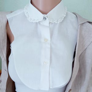 Puede incluir: Un cuello desmontable blanco con un borde de encaje festoneado y un pequeño botón decorativo. El cuello se lleva sobre una camiseta blanca y debajo de una blazer beige. El cuello añade un toque vintage a cualquier atuendo.