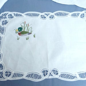Puede incluir: Un camino de mesa de encaje blanco con una imagen bordada de un pato en un estanque. El encaje tiene un borde festoneado.