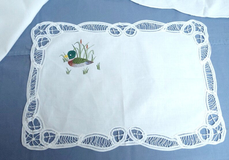 Puede incluir: Un camino de mesa de encaje blanco con una imagen bordada de un pato en un estanque. El encaje tiene un borde festoneado.