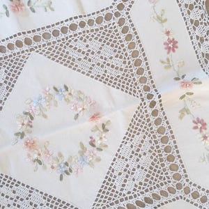 Peut inclure: Une nappe blanche ornée de bordures et de panneaux en dentelle au crochet complexes. Le tissu est décoré de couronnes florales brodées et de motifs de vignes dans des tons doux de rose, bleu et vert, mettant en valeur un travail minutieux.