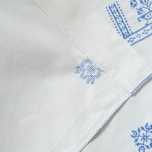 Puede incluir: Textil de lino blanco con patrones florales bordados en azul. El bordado es detallado y aparece en los bordes y esquinas de la tela. La tela est&aacute; doblada, mostrando la textura y el dise&ntilde;o.
