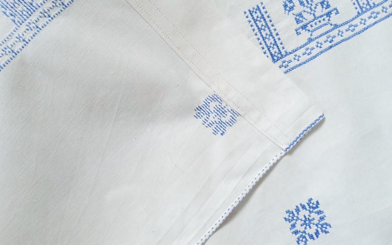 Puede incluir: Textil de lino blanco con patrones florales bordados en azul. El bordado es detallado y aparece en los bordes y esquinas de la tela. La tela est&aacute; doblada, mostrando la textura y el dise&ntilde;o.
