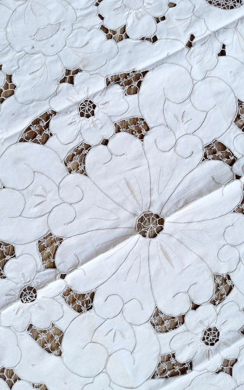 May include: White cotton tablecloth with intricate embroidered floral designs. The tablecloth features a delicate lace pattern and a raised, textured floral motif.