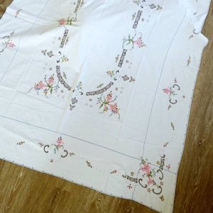 Könnte beinhalten: Weiße Tischdecke mit einem zarten floralen Stickmuster in Rosa, Grün und Braun. Die Stickerei zeigt ein sich wiederholendes Muster aus Blumen und Blättern am Rand und in der Mitte.