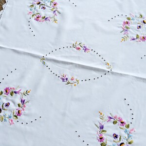 Peut inclure: Nappe blanche avec une bordure en crochet délicate et des motifs floraux brodés dans des tons de rose, violet et bleu.