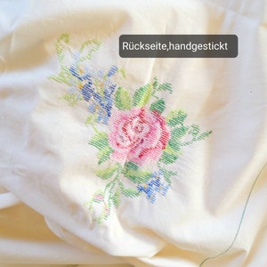 May include: White fabric with a hand-stitched floral design featuring a pink rose, blue flowers, and green leaves. The text "R&uuml;ckseite, handgestickt" is visible in the upper right corner.