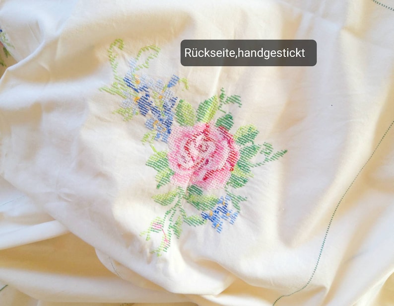 May include: White fabric with a hand-stitched floral design featuring a pink rose, blue flowers, and green leaves. The text "R&uuml;ckseite, handgestickt" is visible in the upper right corner.