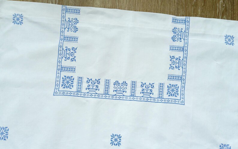 Puede incluir: Tela blanca con un dise&ntilde;o floral bordado en azul. El dise&ntilde;o incluye bordes y patrones de esquina con motivos florales. La tela es probablemente un mantel o un textil decorativo.