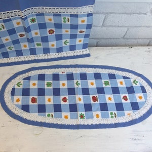 Könnte beinhalten: Ein blauer und weißer karierter ovaler Tischläufer mit einem weißen Häkelrand. Der Läufer hat ein sich wiederholendes Muster aus kleinen roten Äpfeln, gelben Blumen und grünen Blättern.