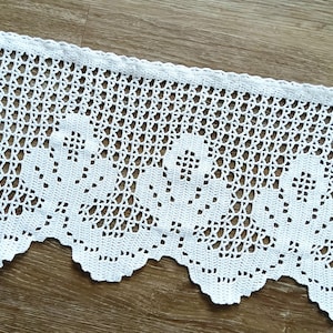 Op de afbeelding: Een stuk witte, gehaakte kanten rand met een geschulpte rand. Het kant heeft een open, bloemachtig patroon. De rand wordt getoond op een houten oppervlak, dat de textuur en het ontwerp laat zien. Deze decoratieve rand is geschikt voor diverse knutselprojecten.