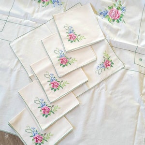 May include: A set of white linen napkins with green embroidered floral designs featuring pink roses. The napkins are arranged on a larger white tablecloth with a similar floral design.