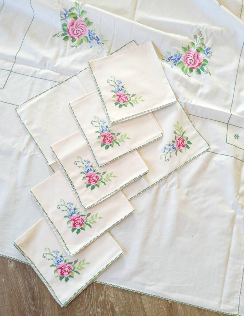 May include: A set of white linen napkins with green embroidered floral designs featuring pink roses. The napkins are arranged on a larger white tablecloth with a similar floral design.