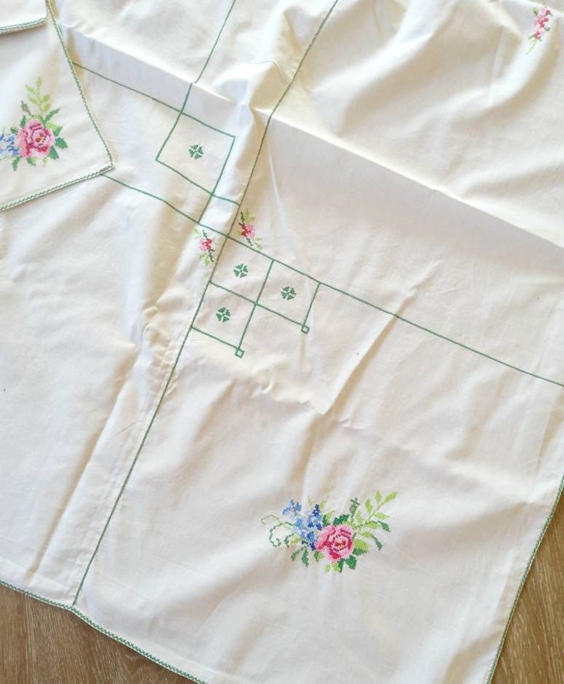 May include: White tablecloth with green embroidered floral designs. The tablecloth has a delicate green border and features a floral pattern with pink, blue, and green flowers.