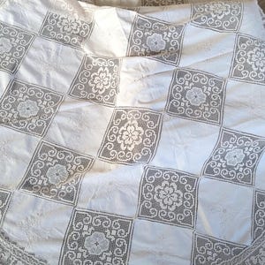 Puede incluir: Un mantel de crochet blanco con un patrón cuadrado y un borde festoneado. El mantel está hecho de encaje delicado y tiene un aspecto vintage.