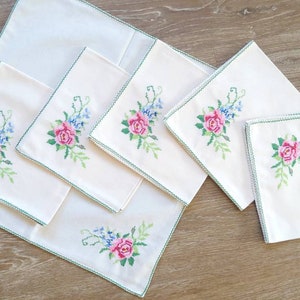May include: Set of six white linen napkins with green embroidered floral designs featuring pink roses and blue flowers.