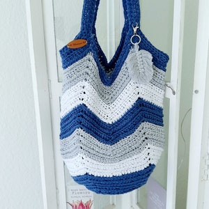 May include: A blue, white, and gray crocheted tote bag with a chevron pattern. The bag has a brown leather tag that says "Handmade" and a gray tassel attached to the handle.