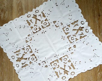 Puede incluir: Un mantel cuadrado de tela blanca con intrincados bordados calados. El diseño muestra motivos florales, hojas y pequeños ojales circulares en el centro y a lo largo de los bordes festoneados, creando un delicado patrón de calados.
