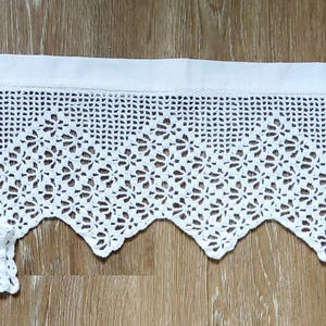 Peut inclure: Une bordure en dentelle crochetée blanche avec un bord festonné. La dentelle présente un motif floral et est fixée à un en-tête en tissu blanc. Plusieurs morceaux de dentelle assortie pliés sont visibles sur le côté gauche, le tout sur une surface en bois.